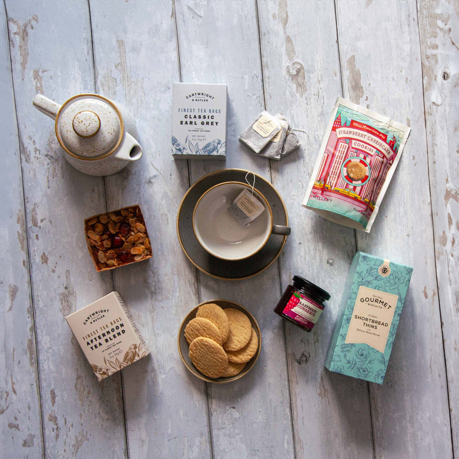 The contents of the Afternoon Tea Hamper by Spicers of Hythe laid out in a grid on a wooden table top including shortbread biscuits, cookies, teabags & more